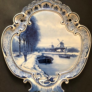 May include: A blue and white decorative ceramic wall plate featuring a winter landscape scene. The plate depicts a river, windmills, and figures, framed by an ornate border. The plate is approximately 12 inches wide.
