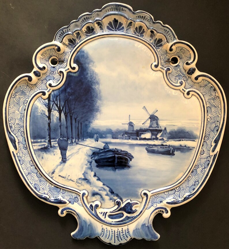 May include: A blue and white decorative ceramic wall plate featuring a winter landscape scene. The plate depicts a river, windmills, and figures, framed by an ornate border. The plate is approximately 12 inches wide.