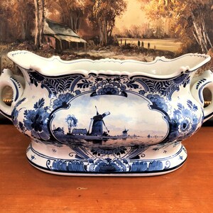 May include: A large, white ceramic planter with blue and white Delftware designs. The planter features a scene of a Dutch windmill and a canal with boats. The planter has two handles and a scalloped rim.