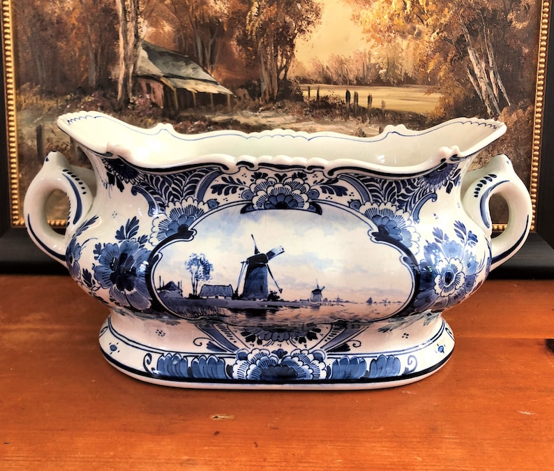 May include: A large, white ceramic planter with blue and white Delftware designs. The planter features a scene of a Dutch windmill and a canal with boats. The planter has two handles and a scalloped rim.