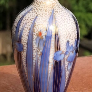 May include: A small, white porcelain vase with a gold and blue floral design. The vase has a wide mouth and a narrow neck, and it sits on a silver base with a clover design.