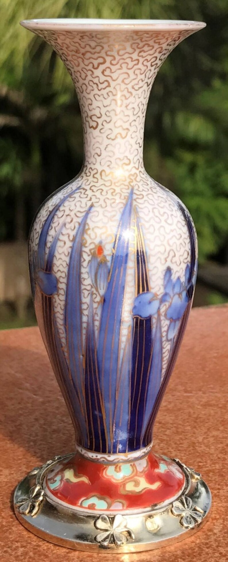 May include: A small, white porcelain vase with a gold and blue floral design. The vase has a wide mouth and a narrow neck, and it sits on a silver base with a clover design.