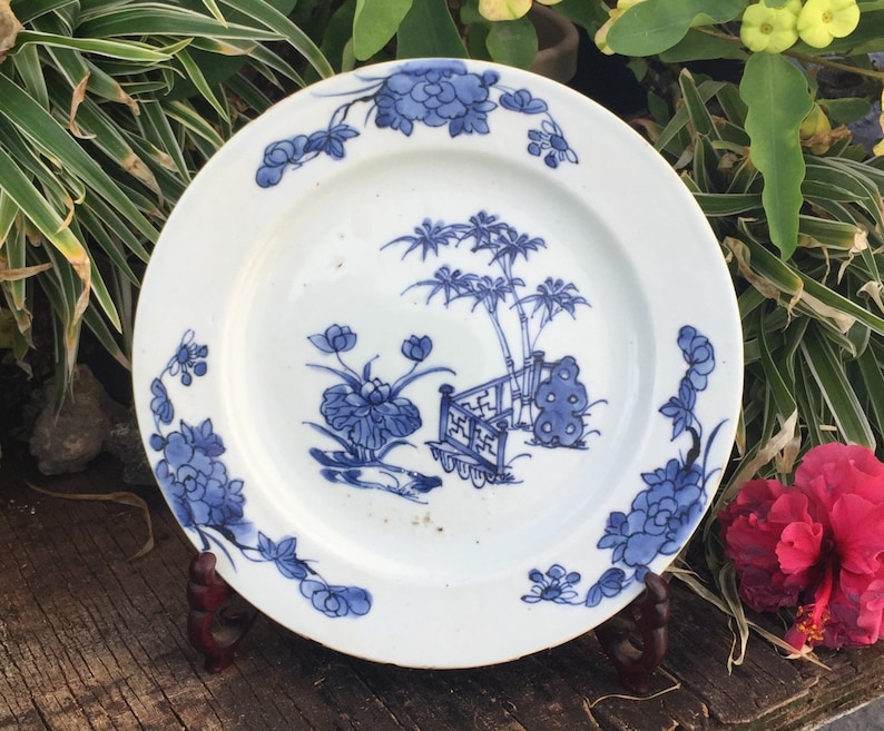 Op de afbeelding: Een wit porseleinen bord met een blauw en wit bloemenmotief. Het bord toont een sc&egrave;ne met een bamboebos, een hek en bloemen. Het bord staat op een houten standaard.