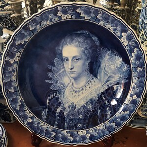 Pu&ograve; includere: Un piatto in maiolica di Delft blu e bianco con un ritratto di una donna con un colletto di pizzo e una collana di perle. Il piatto ha un bordo a festoni ed &egrave; decorato con motivi floreali.