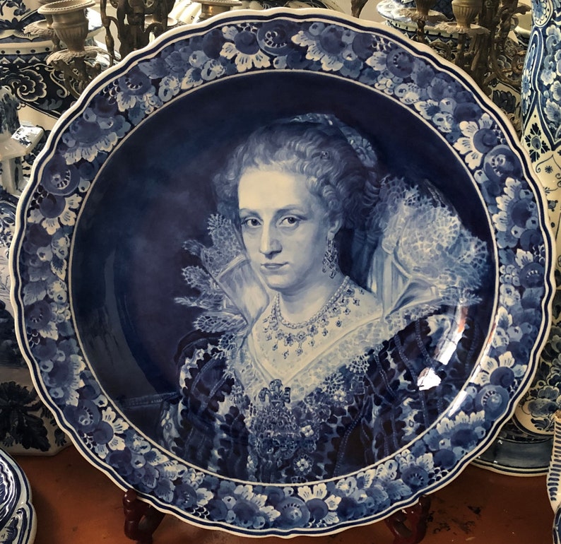 Pu&ograve; includere: Un piatto in maiolica di Delft blu e bianco con un ritratto di una donna con un colletto di pizzo e una collana di perle. Il piatto ha un bordo a festoni ed &egrave; decorato con motivi floreali.