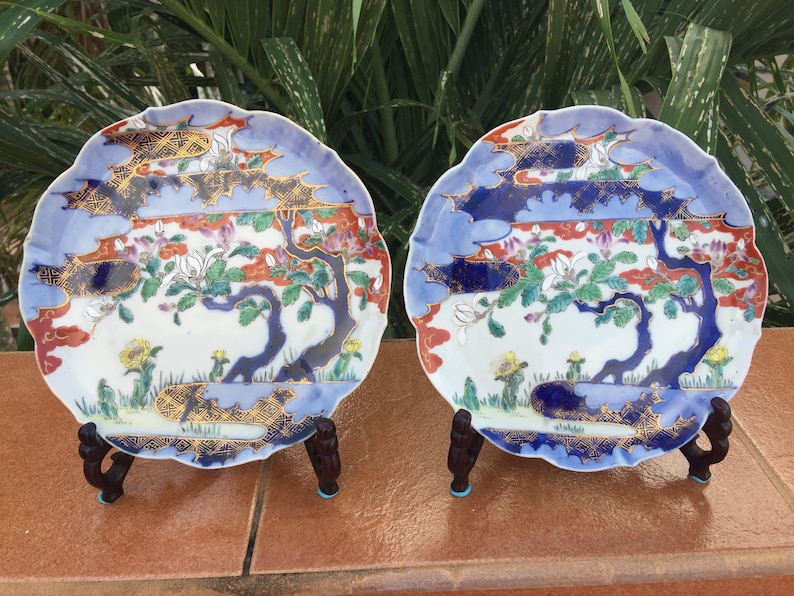 May include: Two decorative ceramic plates with scalloped edges. Each plate features a detailed landscape scene with a blue sky, trees, and floral elements. The plates are supported by dark wooden stands, showcasing an Asian-inspired design.