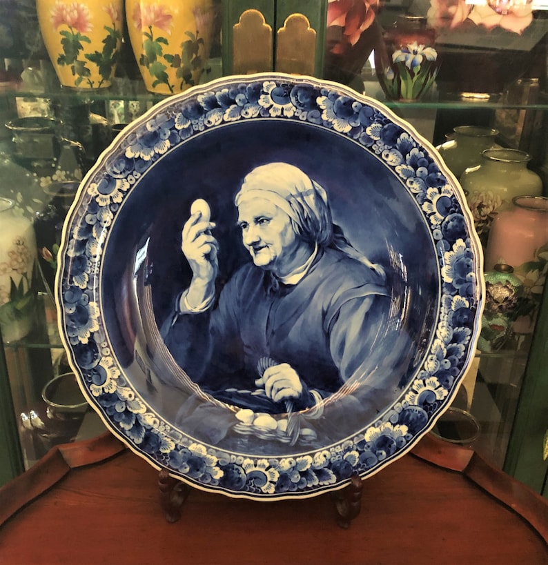 Puede incluir: Un plato de Delftware azul y blanco con un borde elevado y un dise&ntilde;o floral. El centro del plato presenta a una mujer sosteniendo un huevo en su mano.