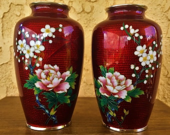 May include: Two red decorative vases with floral designs. Each vase features a large peony and smaller white flowers on branches. The vases have a silver rim at the top and bottom, and a textured surface.