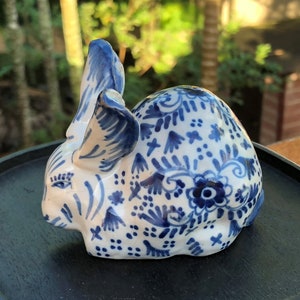 Puede incluir: Figura de conejo de cer&aacute;mica en blanco y azul. El conejo est&aacute; decorado con patrones florales azules y tiene orejas erguidas. La figura est&aacute; sobre una bandeja negra, con un fondo verde borroso.