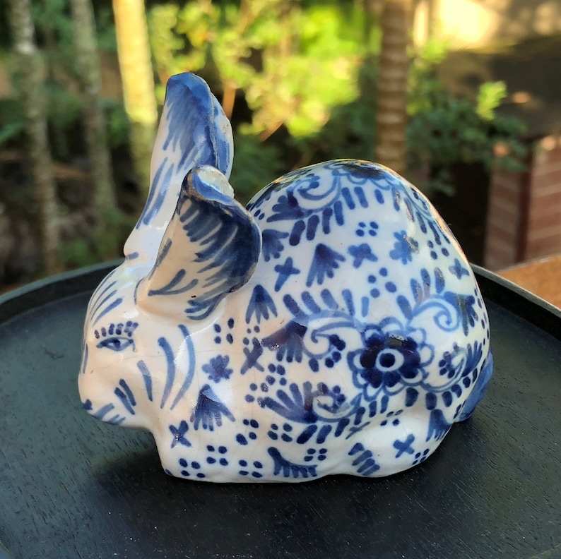 Puede incluir: Figura de conejo de cer&aacute;mica en blanco y azul. El conejo est&aacute; decorado con patrones florales azules y tiene orejas erguidas. La figura est&aacute; sobre una bandeja negra, con un fondo verde borroso.