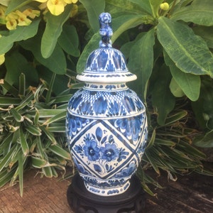 May include: A blue and white ceramic ginger jar featuring intricate hand-painted floral motifs and geometric diamond patterns. The jar includes a matching lid topped with a small sculpted finial and rests on a dark circular wooden display stand.
