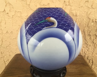 May include: A decorative, spherical vase with a crane design. The vase features a white base with blue accents and a blue wave pattern at the top. The crane has a red head and a long green beak. The vase is displayed on a black stand.