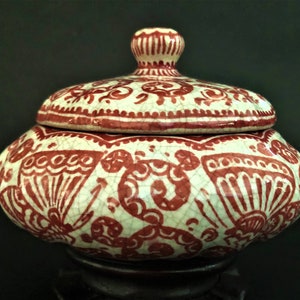 May include: A white ceramic jar with a red floral design and a lid. The jar is on a wooden stand.
