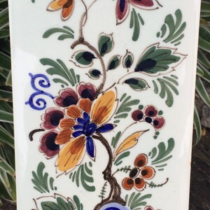 K&ouml;nnte beinhalten: Eine wei&szlig;e Keramikfliese mit handbemaltem Blumendesign. Das Design zeigt blaue, gelbe, orange, gr&uuml;ne und braune Blumen und Bl&auml;tter.