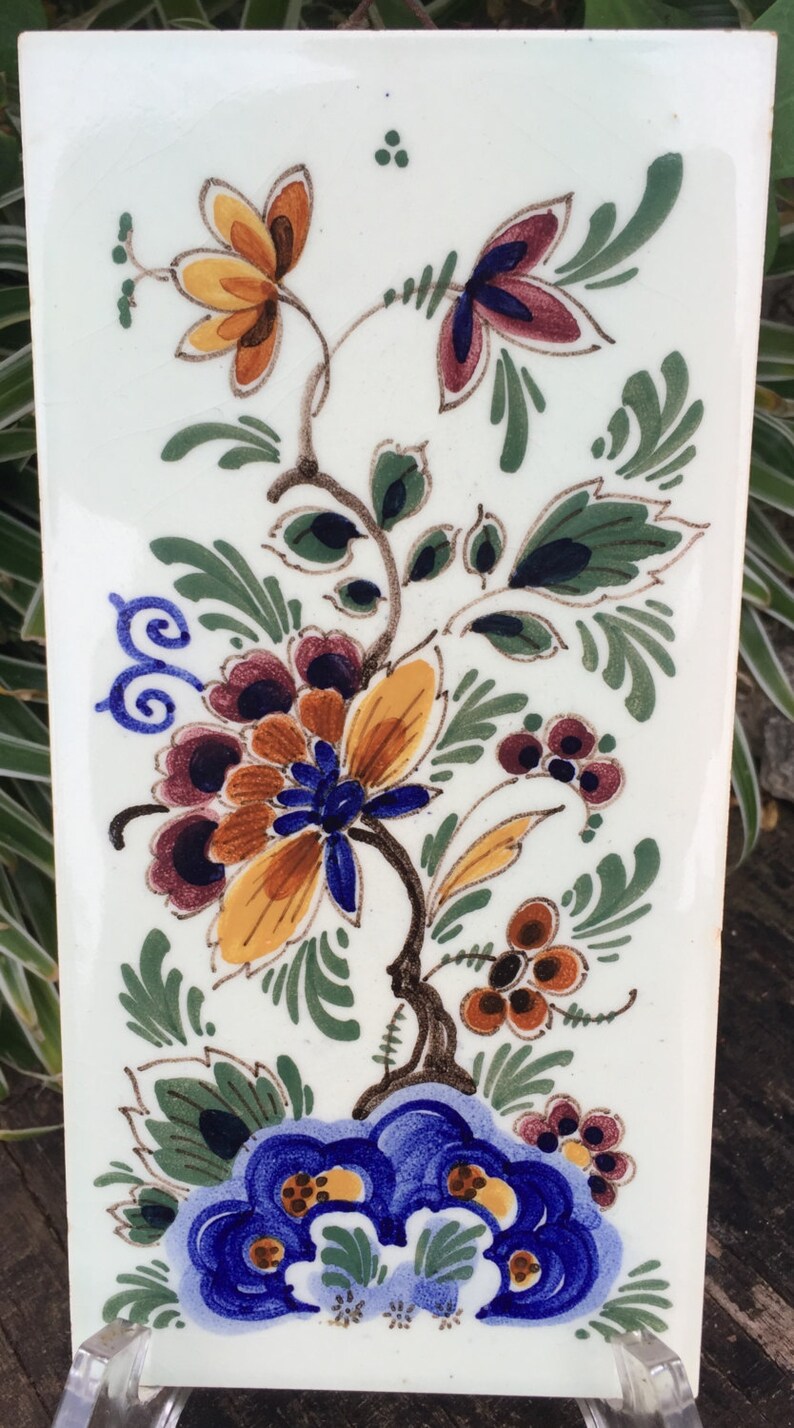 K&ouml;nnte beinhalten: Eine wei&szlig;e Keramikfliese mit handbemaltem Blumendesign. Das Design zeigt blaue, gelbe, orange, gr&uuml;ne und braune Blumen und Bl&auml;tter.