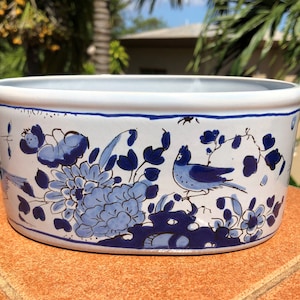 Op de afbeelding: Witte en blauwe keramische bloempot met een bloemen- en vogelontwerp. De bloempot heeft een rechthoekige vorm met afgeronde hoeken. Het ontwerp toont blauwe bloemen en vogels op een witte achtergrond.