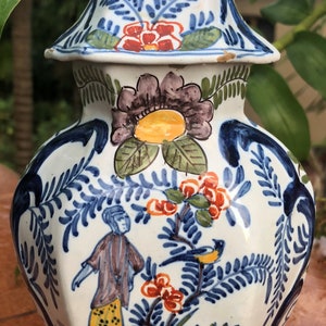 Puede incluir: Jarr&oacute;n de cer&aacute;mica pintado a mano con tapa, con dise&ntilde;os florales y figurativos en azul, rojo, amarillo y marr&oacute;n. El jarr&oacute;n tiene forma hexagonal y un remate decorativo. La altura total es de unos 30 cm.