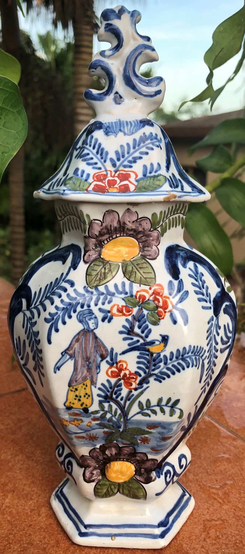 Puede incluir: Jarr&oacute;n de cer&aacute;mica pintado a mano con tapa, con dise&ntilde;os florales y figurativos en azul, rojo, amarillo y marr&oacute;n. El jarr&oacute;n tiene forma hexagonal y un remate decorativo. La altura total es de unos 30 cm.