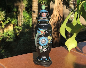 May include: A black decorative vase with a lid, featuring a colorful bird and floral design. The vase has a green finial on top and sits on a small, dark wooden base. The design includes blue, red, and yellow accents.