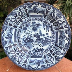 Puede incluir: Un plato grande de cer&aacute;mica azul y blanco con un borde decorativo y una escena central que representa una figura en un paisaje. El plato tiene un esmalte craquelado y un borde dorado.