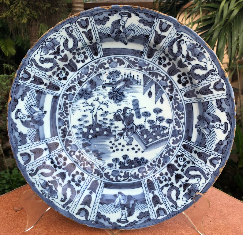 Puede incluir: Un plato grande de cer&aacute;mica azul y blanco con un borde decorativo y una escena central que representa una figura en un paisaje. El plato tiene un esmalte craquelado y un borde dorado.