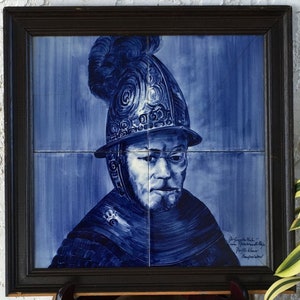 May include: A blue and white ceramic tile depicting a man in a helmet with a plume. The tile is framed in a dark wood frame. The tile is signed "De Gouden Hoede -  Amsterdam" and "P.J. de Kleijn - Handgeschilderd".