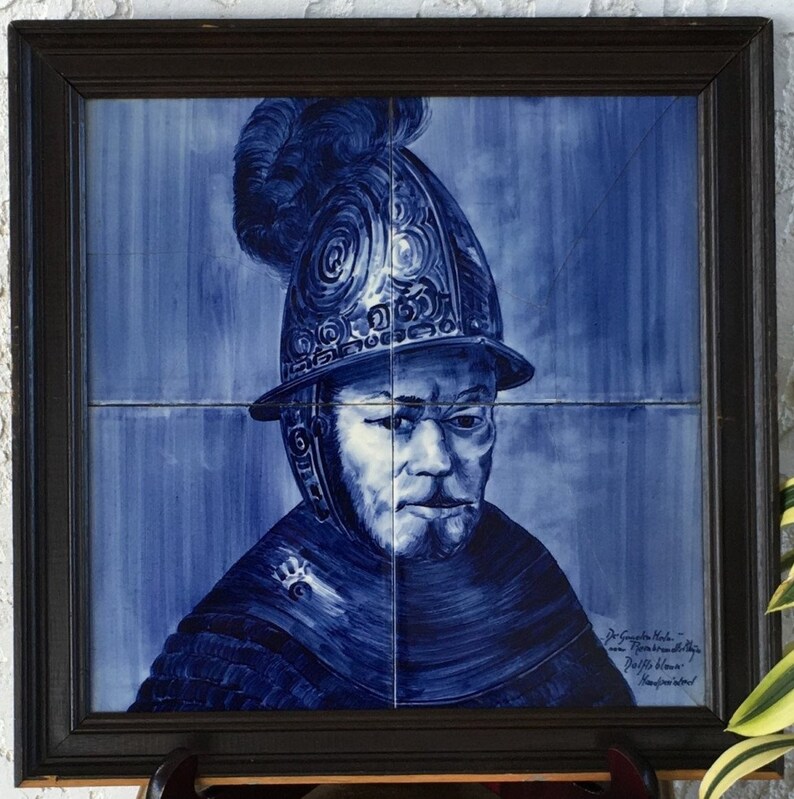 May include: A blue and white ceramic tile depicting a man in a helmet with a plume. The tile is framed in a dark wood frame. The tile is signed "De Gouden Hoede -  Amsterdam" and "P.J. de Kleijn - Handgeschilderd".