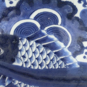 K&ouml;nnte beinhalten: Nahaufnahme eines blau-wei&szlig;en Keramiktellers mit Fischmuster. Der Fisch ist mit detaillierten Schuppen dargestellt und von Wellen- und Wolkenmustern umgeben. Der Teller hat einen dekorativen Rand.