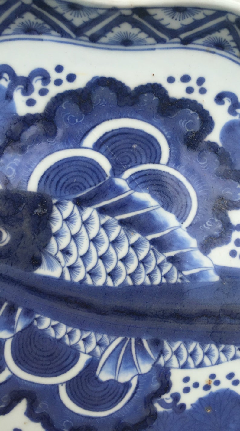 K&ouml;nnte beinhalten: Nahaufnahme eines blau-wei&szlig;en Keramiktellers mit Fischmuster. Der Fisch ist mit detaillierten Schuppen dargestellt und von Wellen- und Wolkenmustern umgeben. Der Teller hat einen dekorativen Rand.