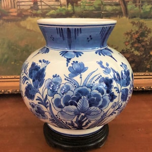 Op de afbeelding: Een witte keramische vaas met een blauw bloemmotief. De vaas heeft een ronde vorm en een uitlopende hals. Hij is versierd met blauwe bloemen en bladeren. De vaas staat op een donkere houten voet. Geschatte hoogte: 20 cm.