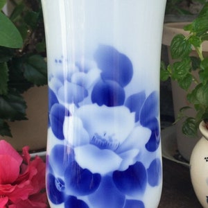 May include: White ceramic vase with a flared top and a round base. The vase is decorated with a blue floral pattern. The vase is sitting on a black stand. A pink flower is visible in the background.