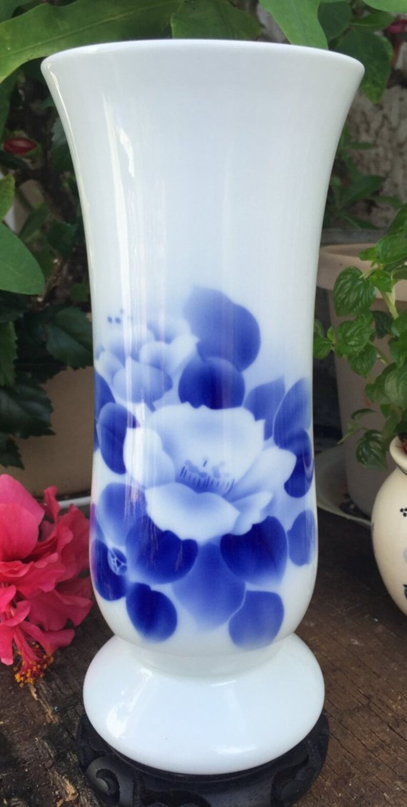 May include: White ceramic vase with a flared top and a round base. The vase is decorated with a blue floral pattern. The vase is sitting on a black stand. A pink flower is visible in the background.