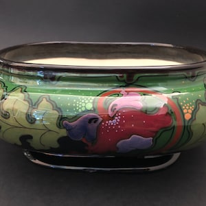 May include: An oval ceramic bowl featuring a glossy green glaze with stylised leaf patterns in shades of light green, dark green, and black. The exterior is decorated with abstract floral motifs in purple, red, and orange, with a dark brown rim and base.