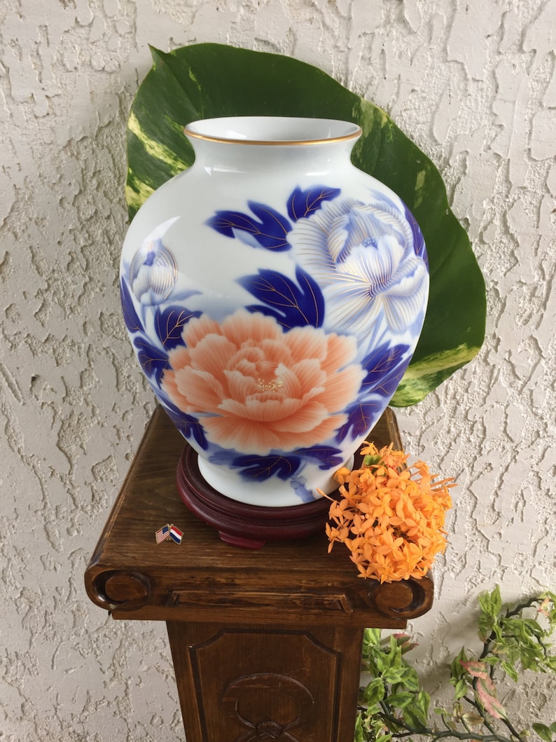 Puede incluir: Un jarr&oacute;n de cer&aacute;mica blanco con borde dorado, con un dise&ntilde;o pintado a mano de una gran peon&iacute;a color melocot&oacute;n y detalles florales azules. El jarr&oacute;n se exhibe sobre un soporte de madera con base roja oscura.