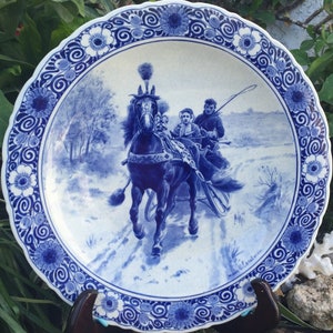 May include: A round, white ceramic plate with a blue floral border and a central illustration of a horse-drawn carriage. The plate features a scalloped edge and a detailed blue and white design, showcasing a historical scene.