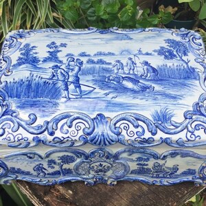 May include: A rectangular, blue and white ceramic box with a hinged lid. The lid features a pastoral scene with figures harvesting crops. The box is decorated with ornate scrollwork and a scalloped edge. The overall design is reminiscent of Delftware.