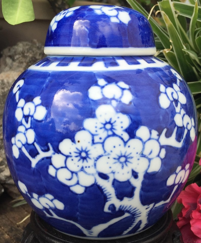 Op de afbeelding: Een kleine blauwe en witte porseleinen pot met een deksel. De pot is versierd met een bloemmotief van witte bloesems op blauwe takken. De pot staat op een houten voet.