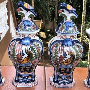 Może przedstawiać: Dwa pasujące wazony ceramiczne i dwa pasujące słoiki ceramiczne z pokrywkami. Słoiki mają ptasią gałkę na pokrywce. Wszystkie elementy są białe z niebieskimi, ż&oacute;łtymi, czerwonymi i zielonymi wzorami kwiatowymi i ptasimi.