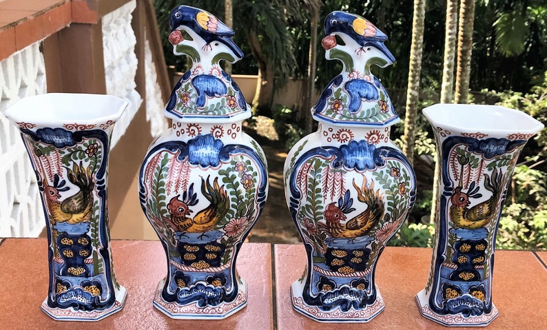 Może przedstawiać: Dwa pasujące wazony ceramiczne i dwa pasujące słoiki ceramiczne z pokrywkami. Słoiki mają ptasią gałkę na pokrywce. Wszystkie elementy są białe z niebieskimi, ż&oacute;łtymi, czerwonymi i zielonymi wzorami kwiatowymi i ptasimi.
