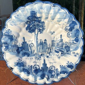 Puede incluir: Un plato de cer&aacute;mica blanca con un dise&ntilde;o pintado a mano en azul y blanco. El plato presenta un &aacute;rbol, dos figuras y un paisaje con flores y follaje. El plato tiene un borde festoneado.