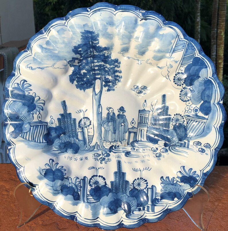 Puede incluir: Un plato de cer&aacute;mica blanca con un dise&ntilde;o pintado a mano en azul y blanco. El plato presenta un &aacute;rbol, dos figuras y un paisaje con flores y follaje. El plato tiene un borde festoneado.