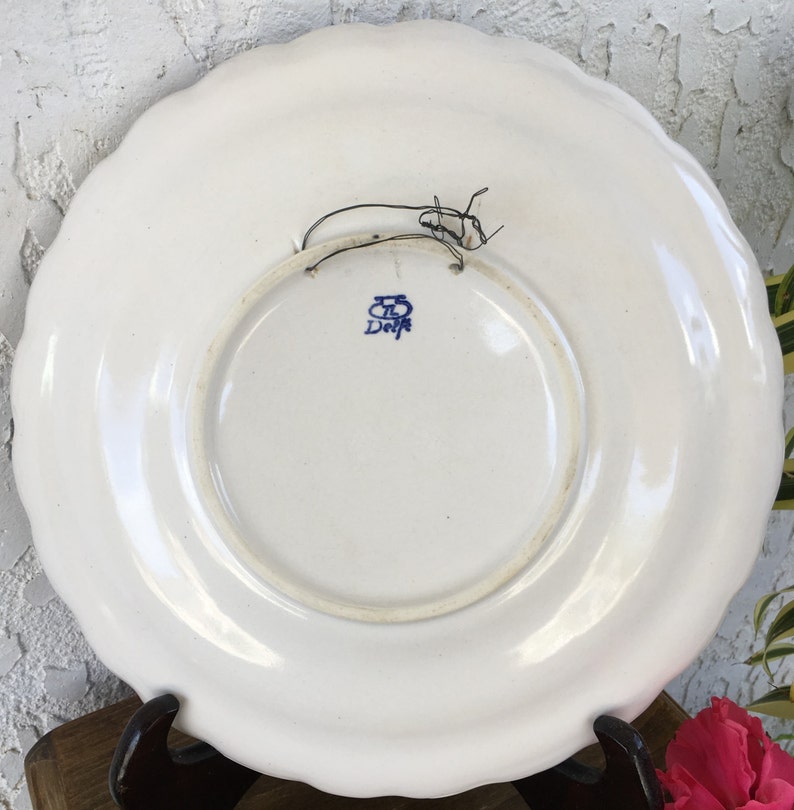 Puede incluir: Plato de cer&aacute;mica blanco con un dise&ntilde;o floral azul y blanco. La parte posterior del plato tiene un sello azul que dice "Delft" con una "B" estilizada encima.