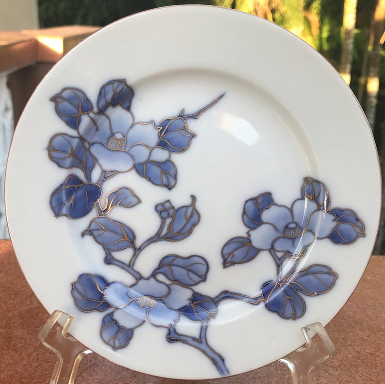 May include: White ceramic plate with a floral design. The plate features blue flowers and leaves with gold accents. The flowers are arranged in a delicate pattern on the plate's surface. The plate has a gold rim.