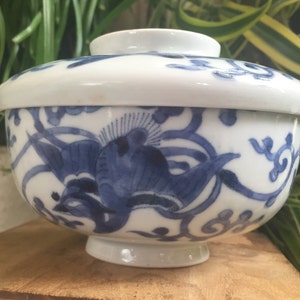 May include: A white ceramic bowl with a lid, featuring a blue floral design. The bowl has a footed base and a rounded shape, with the lid resting on top. The blue design includes stylized birds and swirling patterns.