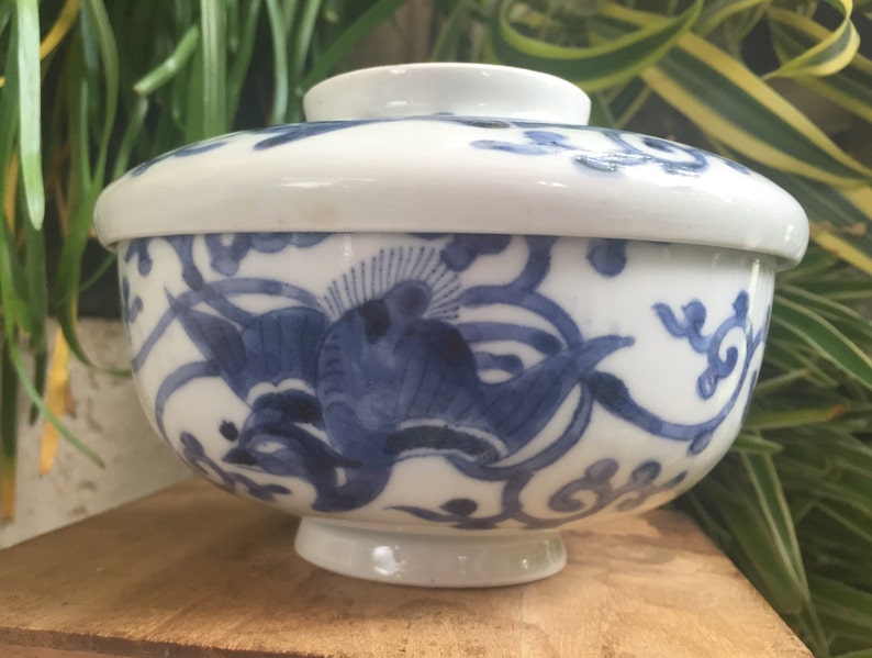 May include: A white ceramic bowl with a lid, featuring a blue floral design. The bowl has a footed base and a rounded shape, with the lid resting on top. The blue design includes stylized birds and swirling patterns.