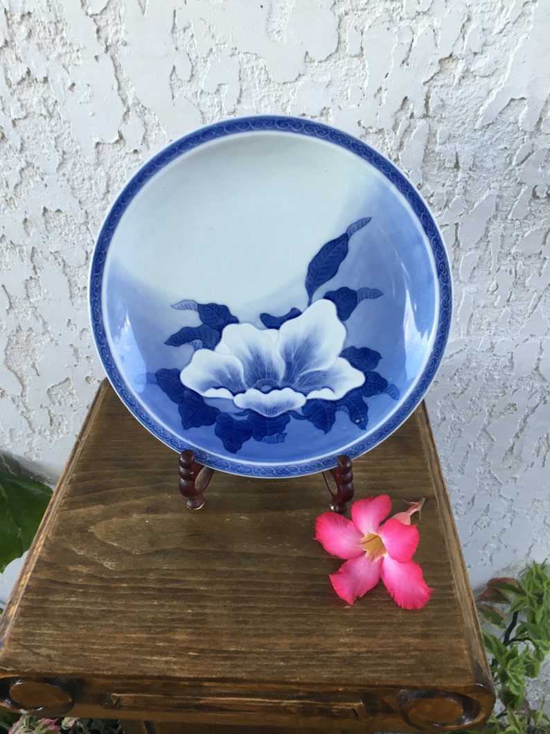 Op de afbeelding: Een ronde keramische plaat in wit en blauw, versierd met een bloemmotief. De plaat heeft een grote witte bloem met blauwe details en een blauwe rand. Hij wordt gepresenteerd op een houten standaard.