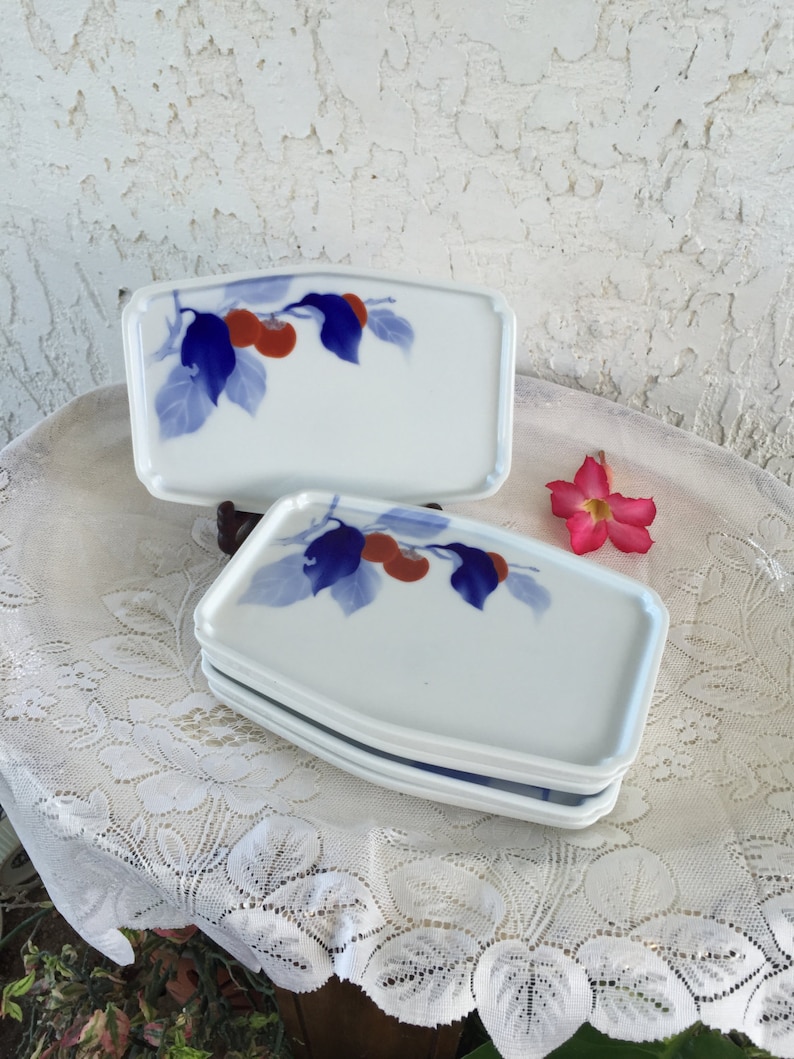 May include: Set of four white ceramic plates with a blue and red floral design. The plates are slightly curved and have a rounded edge.