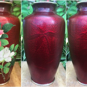 May include: A red decorative vase with a textured surface and a silver rim. The vase is adorned with a floral design featuring pink and white roses with green leaves. A beautiful piece for home decor.