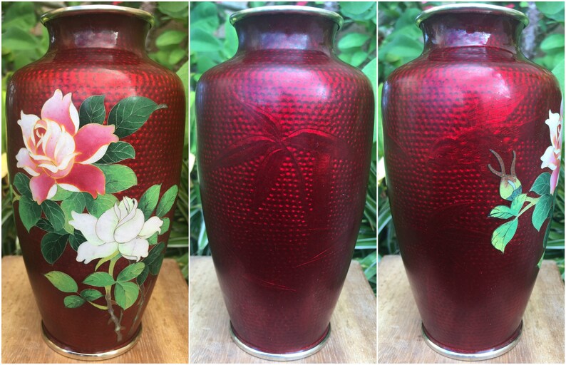 May include: A red decorative vase with a textured surface and a silver rim. The vase is adorned with a floral design featuring pink and white roses with green leaves. A beautiful piece for home decor.