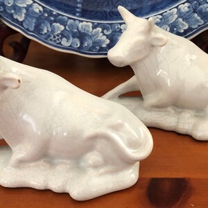 K&ouml;nnte beinhalten: Zwei wei&szlig;e Keramik-Kuhfiguren in entspannter Pose. Die Figuren haben eine Craquel&eacute;-Glasur und ruhen auf einem Sockel. Dekorationsartikel f&uuml;r ein Regal oder einen Tisch.